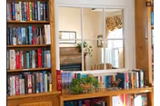 Wooden bookshelf filled with books and a small plant in front of a mirror reflecting a couch and window treatments.