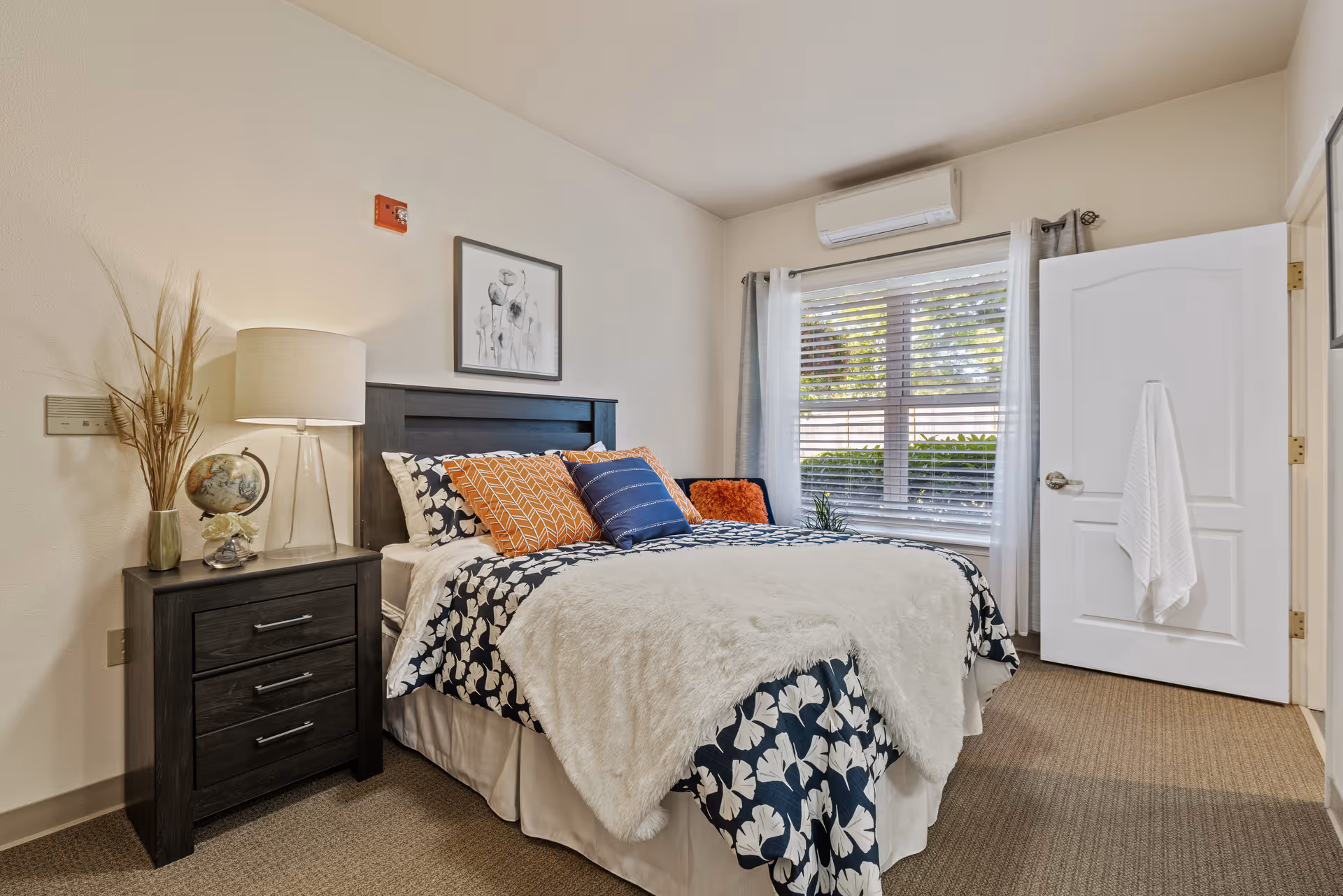 A cozy bedroom with a bed featuring a dark wooden headboard, white and navy floral bedding, and multiple decorative pillows in orange and blue. There is a dark wooden nightstand with a lamp, a globe, and a vase with dried plants. A window with blinds and light curtains lets in natural light, and a white door with a towel hanging on the handle is open. The room has beige carpet and light-colored walls.