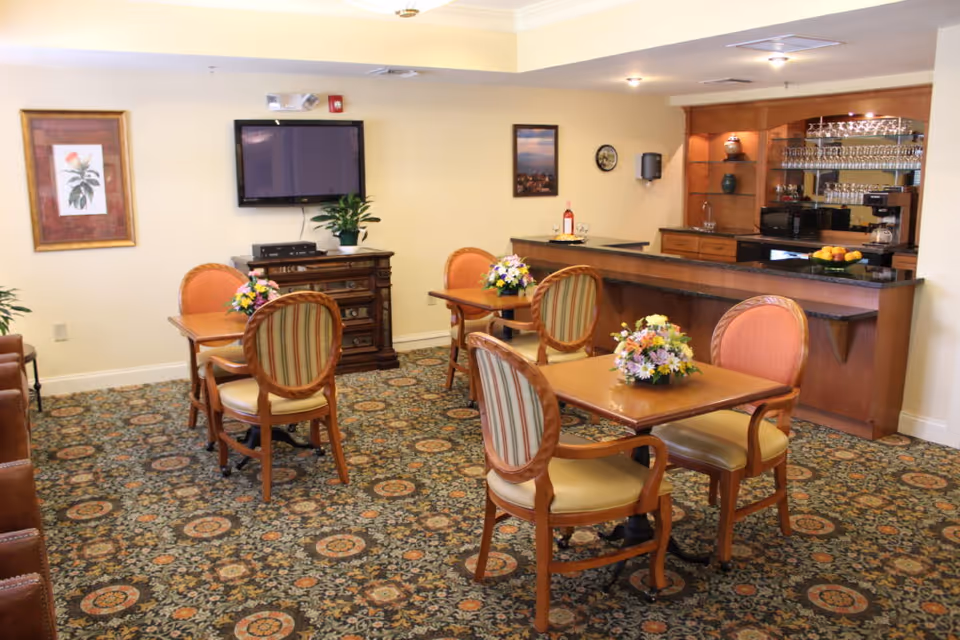A cozy dining area with four wooden tables, each adorned with a floral centerpiece. The room features patterned carpet flooring, a wall-mounted flat-screen TV, framed artwork, and a wooden cabinet with a countertop bar area displaying glassware and a coffee machine.