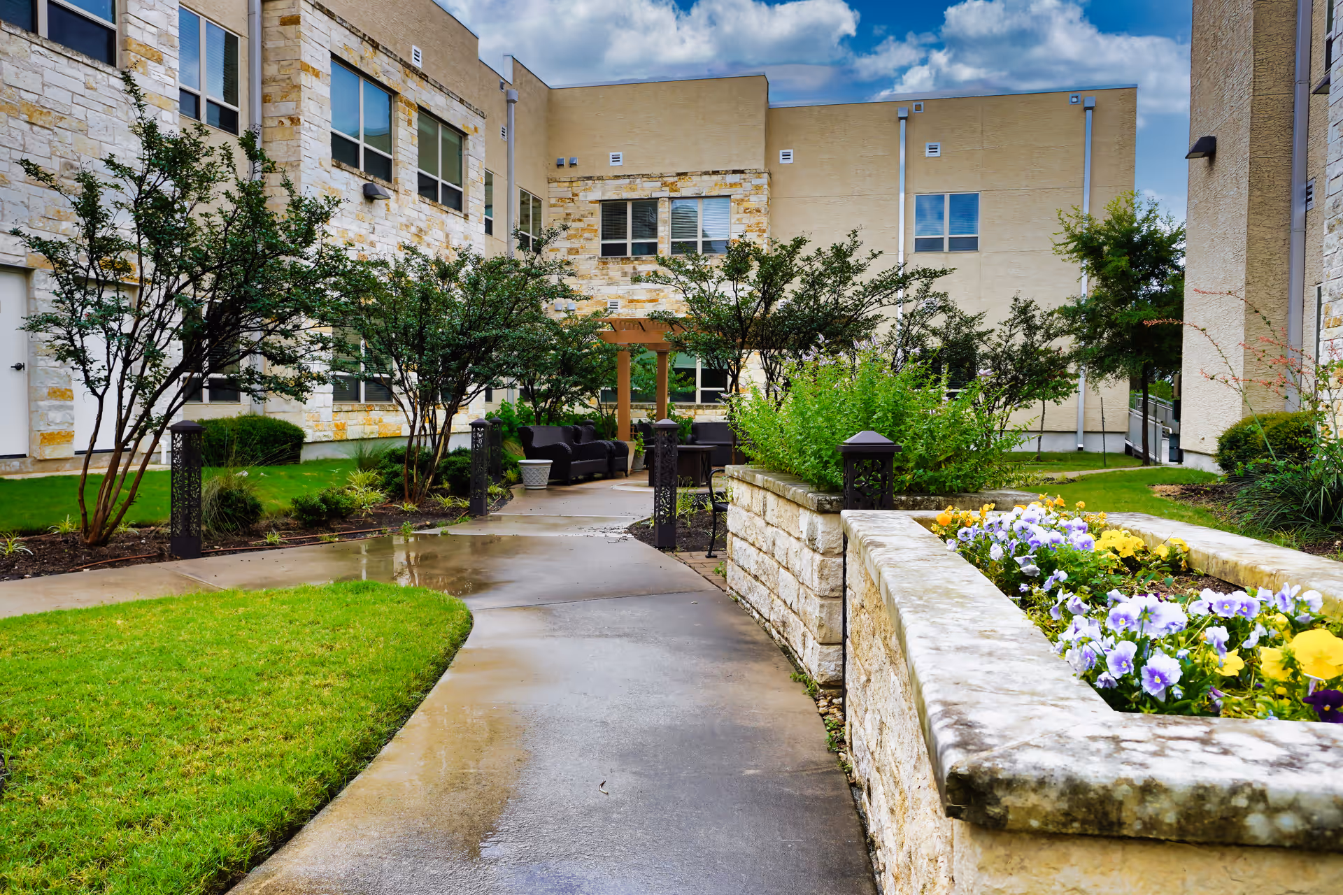Outdoor courtyard with a wet curved walkway, raised stone planters with flowers, shrubs, and seating in front of a multi-story stone-and-stucco building.