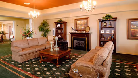 A cozy living room with two patterned sofas facing each other across a wooden coffee table. The room features a fireplace with decorative items on the mantel, two wooden bookshelves with plants and ornaments, a green patterned carpet, and chandeliers hanging from the ceiling. There are framed pictures on the walls and a small round marble-topped side table in the foreground.