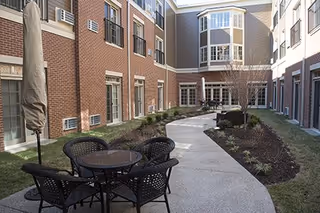 Brick-clad senior living courtyard with a paved walkway, patio table and chairs, and landscaped planting beds.