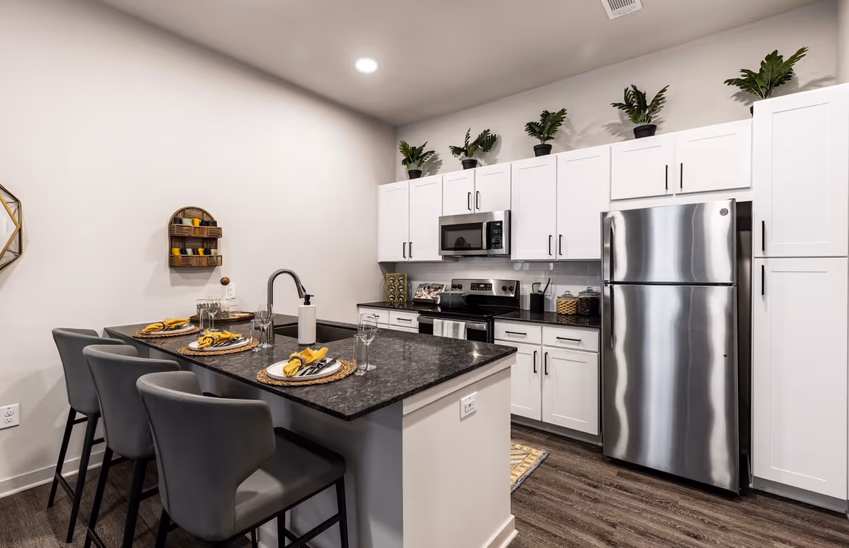 Modern kitchen with white cabinets, stainless steel refrigerator and microwave, black granite countertops, and a kitchen island with four gray chairs set with plates, glasses, and yellow napkins. Several potted plants are placed on top of the cabinets.