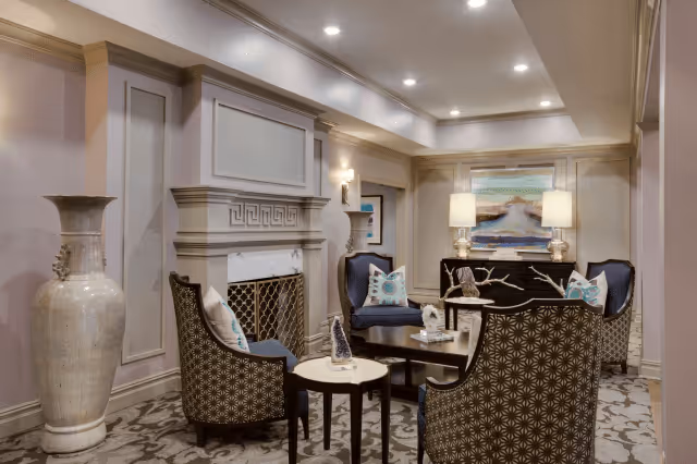 A cozy and elegant living room area with patterned armchairs arranged around a small coffee table. The room features a decorative fireplace with a marble surround, a large ceramic vase, and a sideboard with two table lamps and a landscape painting above it. The walls have detailed molding and the ceiling is fitted with recessed lighting.