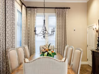 A dining room with a long wooden table surrounded by eight upholstered chairs. A decorative chandelier hangs above the table, and a floral centerpiece is placed in the middle. The room has tall windows with patterned curtains and beige walls.