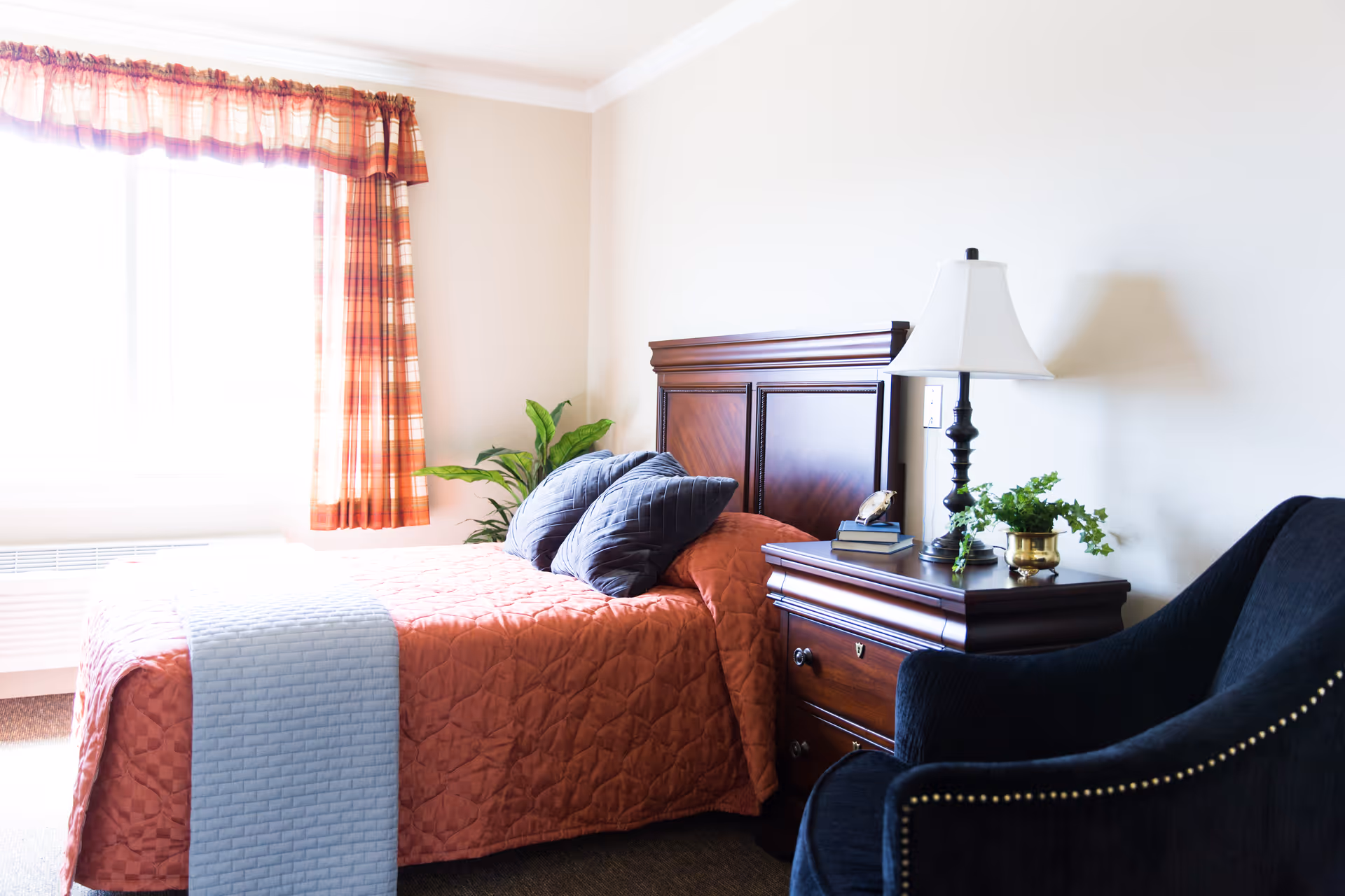 A bright bedroom with a wooden bed frame and an orange quilted bedspread, two gray pillows, a wooden nightstand with a white lamp, books, a clock, and a small potted plant. There is a large window with orange plaid curtains and a dark upholstered armchair with nailhead trim next to the nightstand.