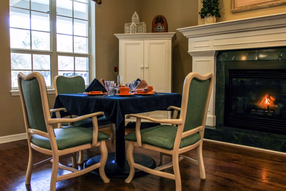 A cozy dining area with a square table covered by a dark blue tablecloth, set with orange plates, glasses, and folded napkins. Four wooden chairs with green cushions surround the table. In the background, there is a white cabinet and a fireplace with a small fire burning. A large window lets in natural light.