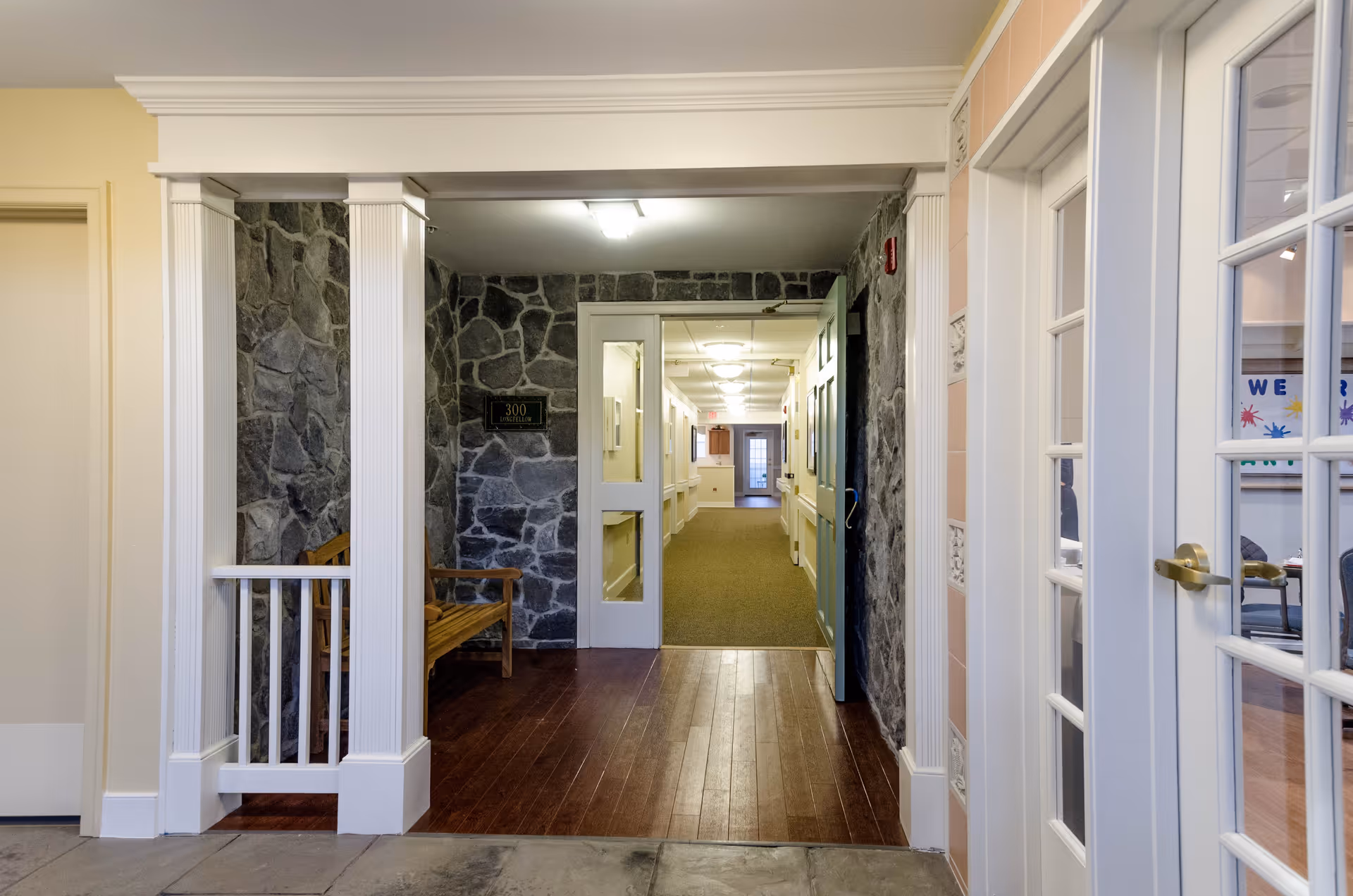 Interior hallway of a senior living facility with stone accent walls, wooden bench, and white columns. The hallway leads to a carpeted corridor with doors on either side. To the right, there are glass-paneled double doors revealing a room with chairs and a bulletin board.