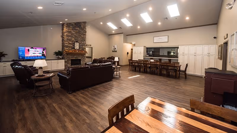 Spacious interior common area with wooden flooring, featuring a seating area with leather sofas and a stone fireplace with a clock above it. A television is mounted on the left wall above white cabinets. To the right, there is a long dining table with chairs and a kitchen area with white cabinetry. The room is well-lit with ceiling lights and skylights.