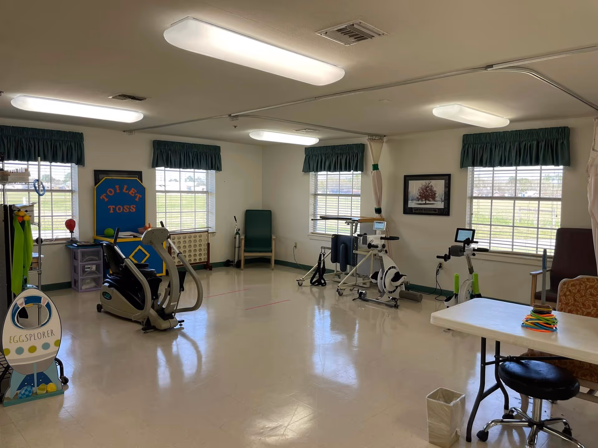 A well-lit activity and exercise room with large windows covered by green valances. The room contains exercise equipment including stationary bikes, a table with ring toss game pieces, and a board labeled 'Toilet Toss'. There are chairs along the walls and a framed picture hanging on one wall.