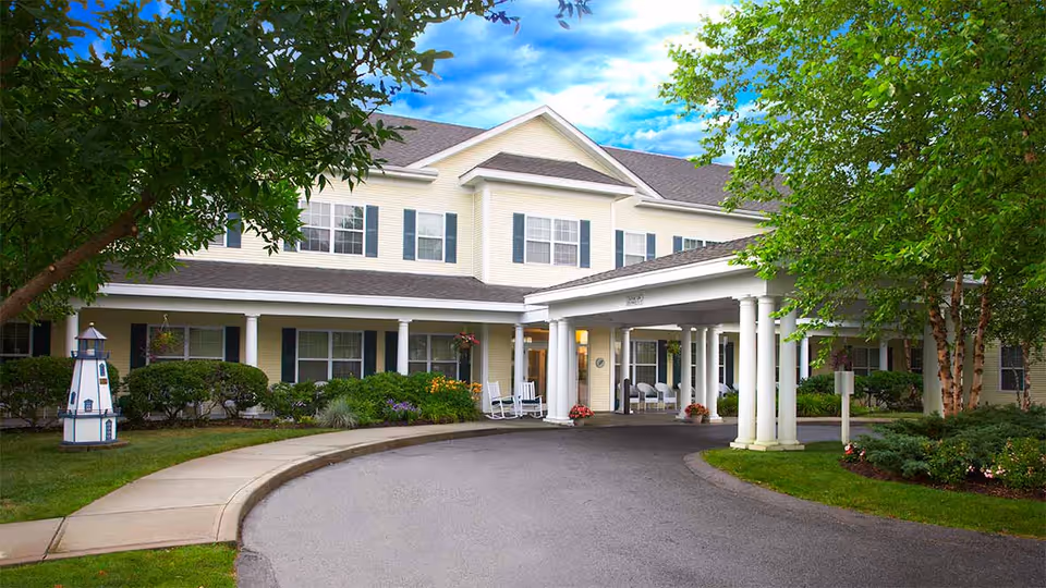 Exterior view of a two-story senior living facility with a covered entrance supported by white columns. The building is light yellow with white trim and dark shutters. There are trees, shrubs, and flowers around the entrance, and a curved driveway leads up to the entrance. The sky is partly cloudy with blue patches.