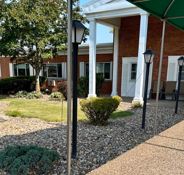 Outdoor view of a senior living facility with a brick building featuring white columns and windows. There is a landscaped area with bushes, trees, and rocks, along with a paved walkway and several black lamp posts. A green canopy is partially visible on the right side.