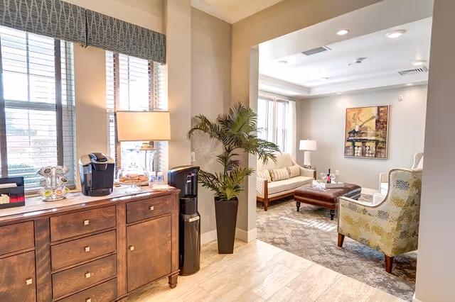 A cozy living room area in a senior living facility with a beige sofa, a patterned armchair, a brown leather ottoman, a floor plant in a black pot, and a wooden sideboard with a coffee maker and lamp. Large windows with blinds allow natural light to fill the space.