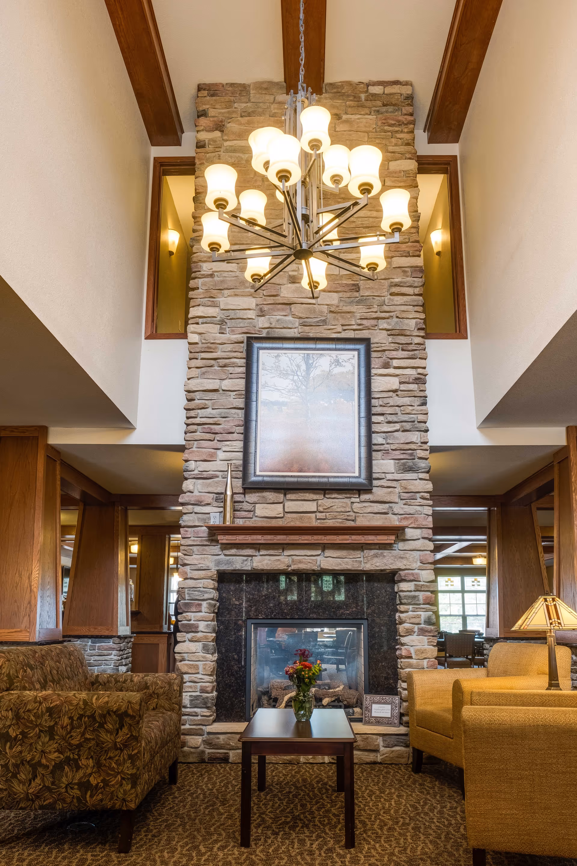 A cozy living room area with a tall stone fireplace as the focal point. Above the fireplace hangs a framed picture, and a modern chandelier with multiple lights is suspended from the ceiling. There are two armchairs, one floral patterned and one solid yellow, positioned on either side of a small wooden coffee table with a vase of flowers. The room features wooden beams on the ceiling and warm lighting.
