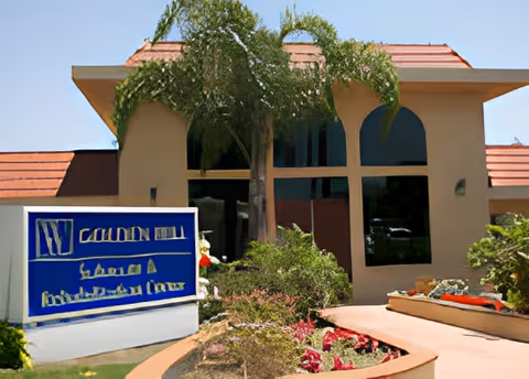 Exterior view of a building with a tan facade and a red-tiled roof, featuring large arched windows. In front of the building is a landscaped area with a curved walkway, plants, flowers, and a palm tree. A blue and white sign is visible near the entrance.