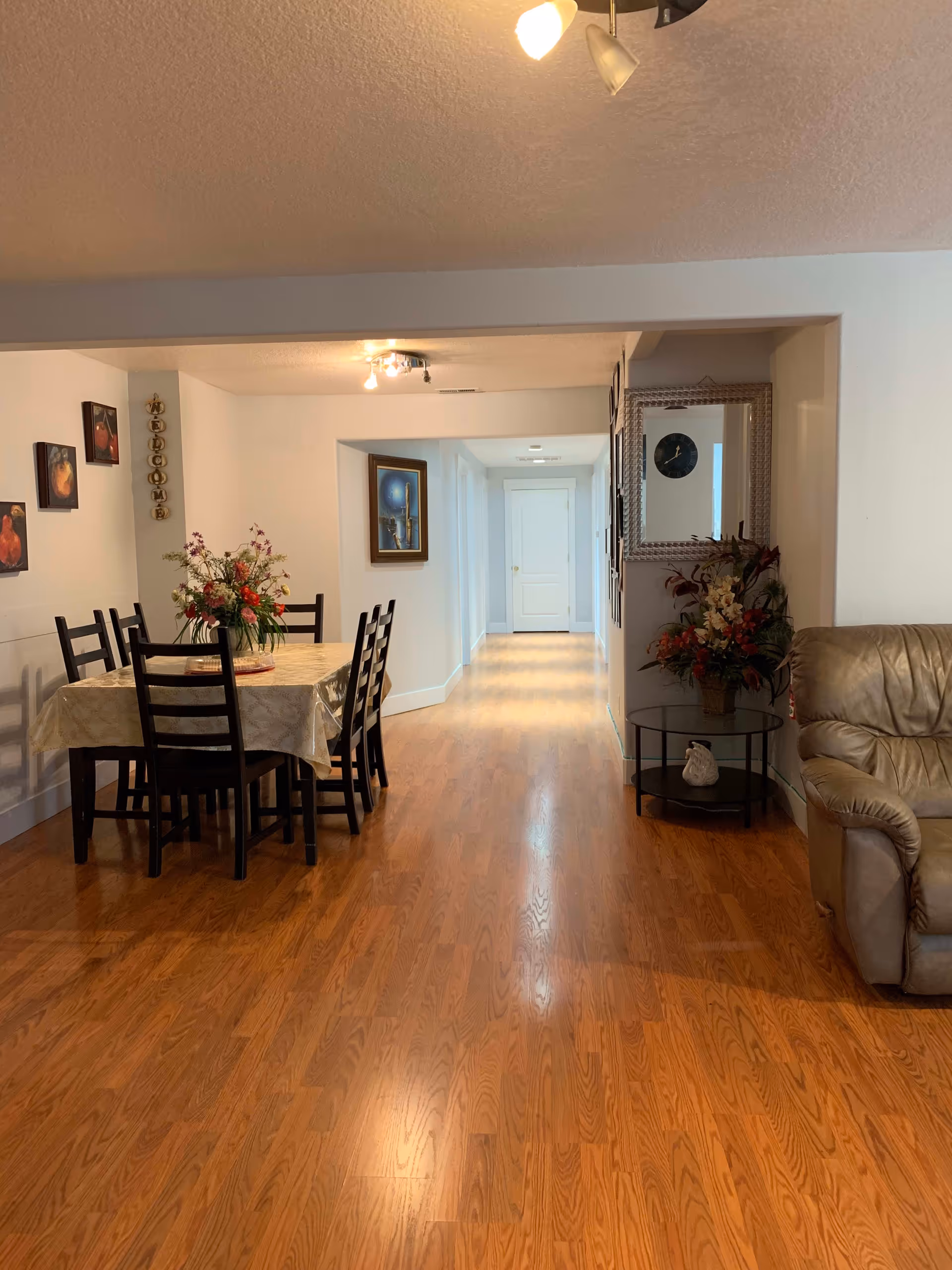 Interior view of a senior living facility showing a dining area with a table covered by a tablecloth and six chairs. A flower arrangement is placed on the table. The room has wooden flooring and white walls decorated with paintings and a vertical welcome sign. To the right, there is a small round glass table with a flower arrangement and a beige leather armchair. A hallway with a white door at the end extends from the dining area.