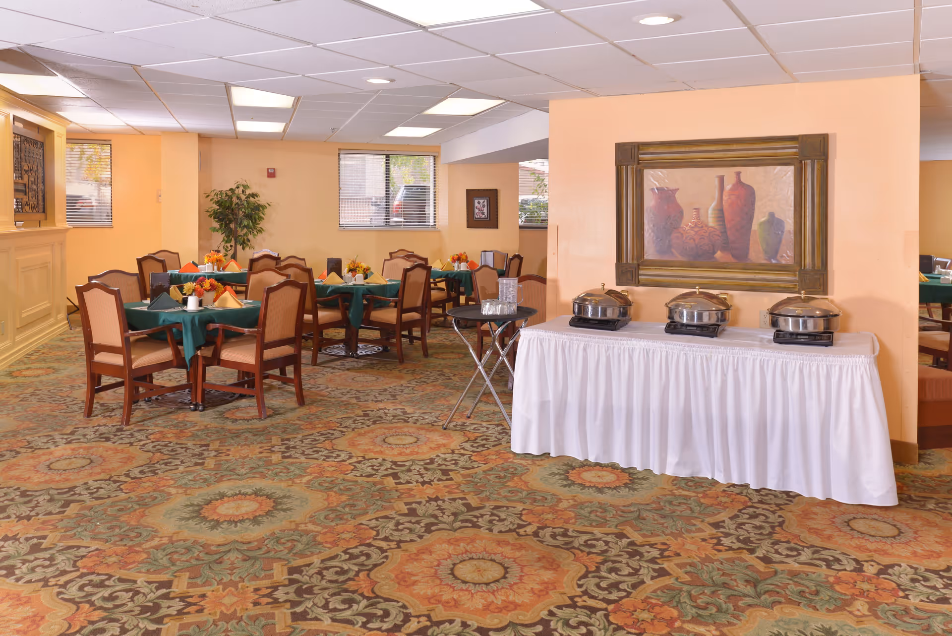 A dining room in a senior living facility with several tables covered with green tablecloths and set with napkins and small floral centerpieces. There is a buffet table with three covered chafing dishes and a small round table with water pitchers and glasses. The room has patterned carpet, beige walls, and a framed painting of vases on the wall.