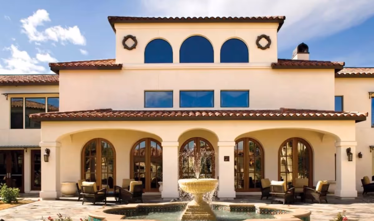 Exterior view of a Mediterranean-style building with arched windows and doors, a tiled roof, and a central water fountain in front. Outdoor seating with cushioned chairs is arranged under the covered patio area.