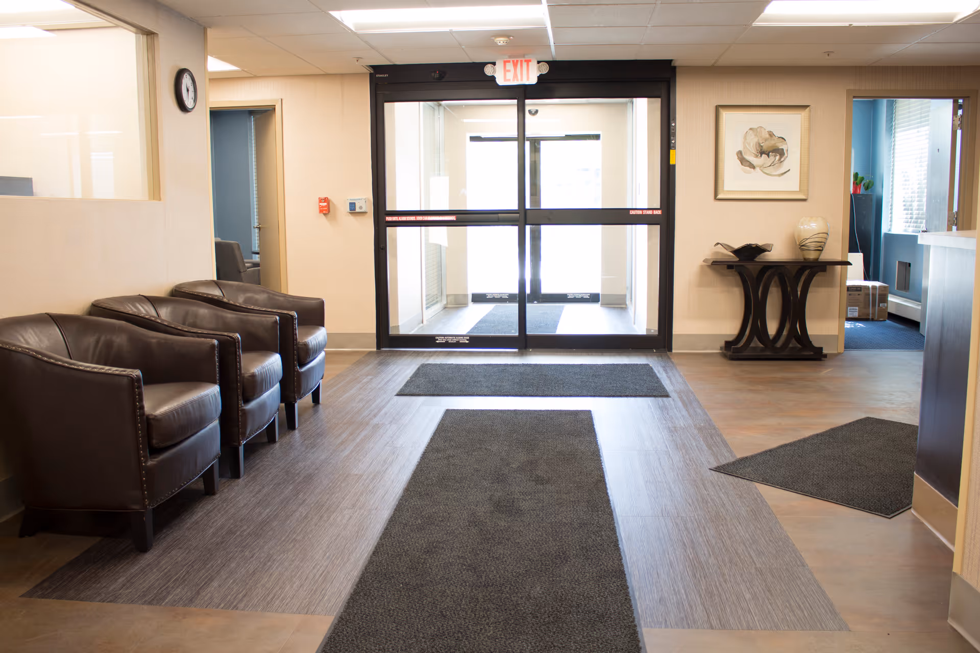 A clean and well-lit waiting area with three brown leather armchairs lined up against the left wall. The floor has wood and tile patterns with dark gray rugs. At the far end, there is a glass automatic door with an exit sign above it. On the right side, there is a small decorative table with a vase and a framed artwork hanging above it. A clock is mounted on the left wall near a window opening.