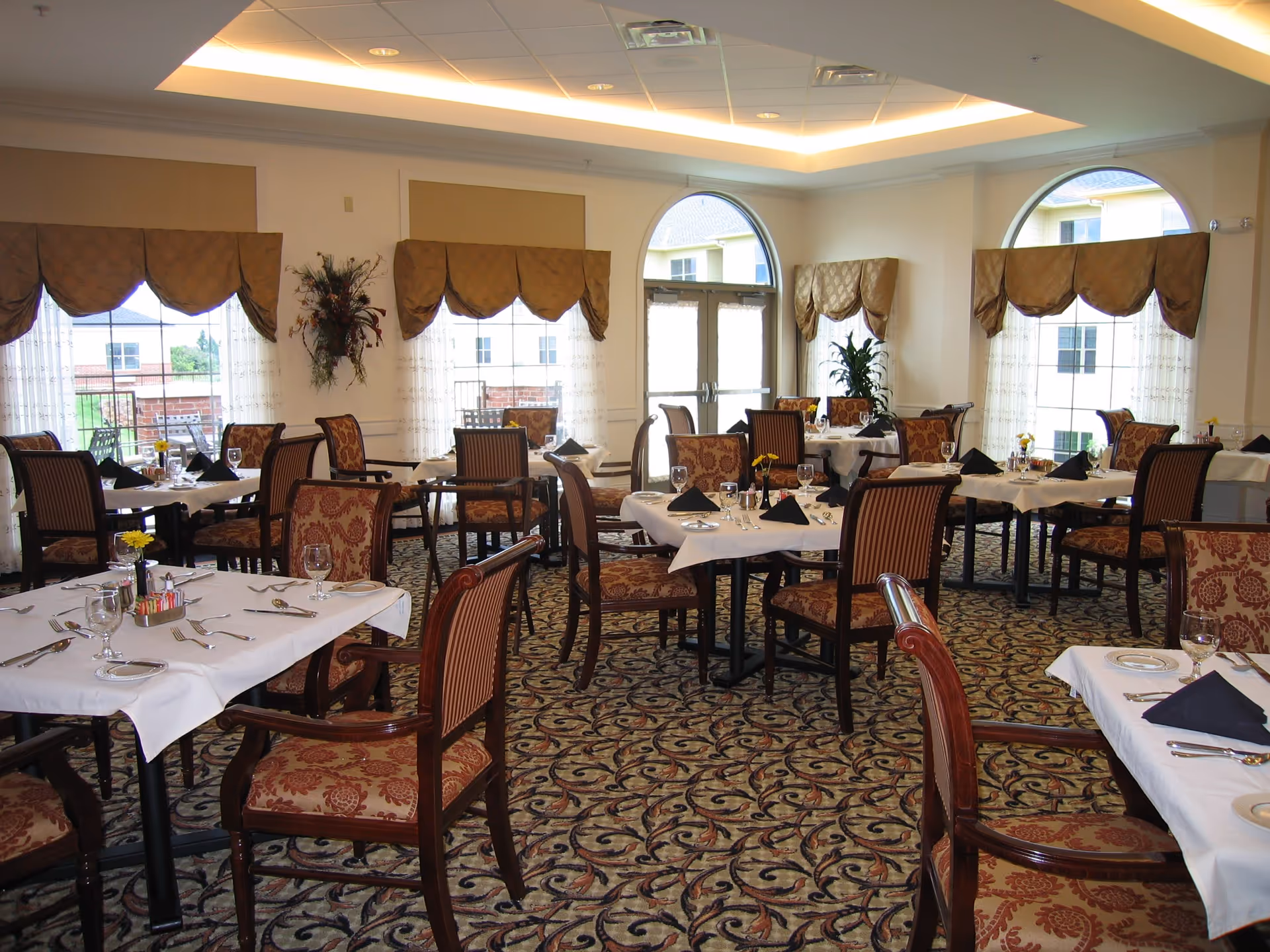 A well-lit dining room with multiple tables covered in white tablecloths, each set with plates, glasses, silverware, and black folded napkins. The chairs have wooden frames with patterned upholstery. Large windows with decorative valances allow natural light to fill the room, and there are plants and floral arrangements on the walls and tables.