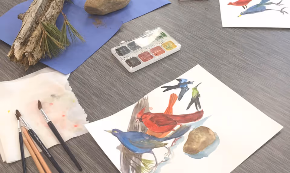 A tabletop with watercolor painting supplies including paintbrushes, a watercolor palette, a piece of tissue paper, and a painting of colorful birds on a branch. There are also natural elements like a piece of bark, pine needles, and a rock placed on blue paper in the background.