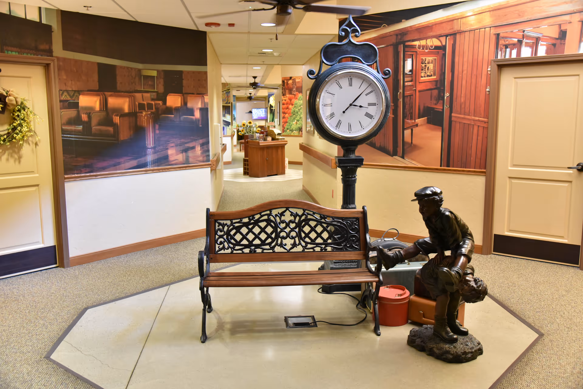 Indoor hallway area with a decorative bench and a vintage-style standing clock. Next to the bench is a bronze statue of a boy putting on a baseball glove. The walls have large murals depicting cozy indoor scenes, and there are two closed doors on either side of the hallway. The floor is carpeted, and the ceiling has recessed lighting and ceiling fans.