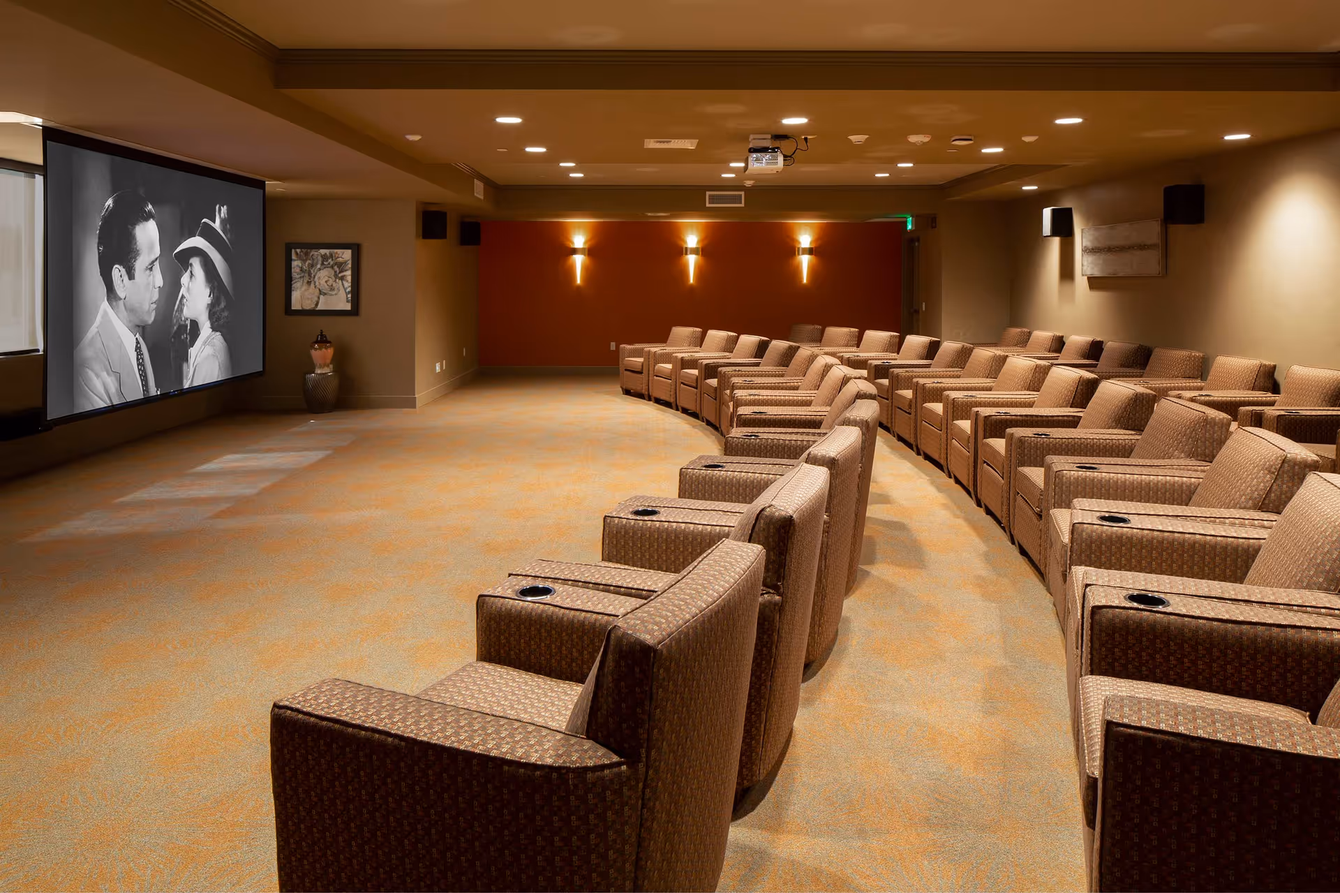 A cozy movie theater room with multiple rows of comfortable brown armchairs arranged in a curved layout facing a large screen showing a black and white movie scene featuring a man and a woman.