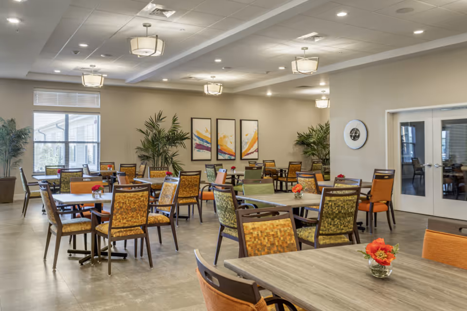 A spacious dining room with multiple tables and chairs arranged neatly. The chairs have colorful upholstery in shades of orange, green, and patterned designs. Each table has a small vase with red flowers. The room has large windows letting in natural light, potted plants, modern ceiling lights, and abstract artwork on the walls. There are double glass doors on one side of the room.