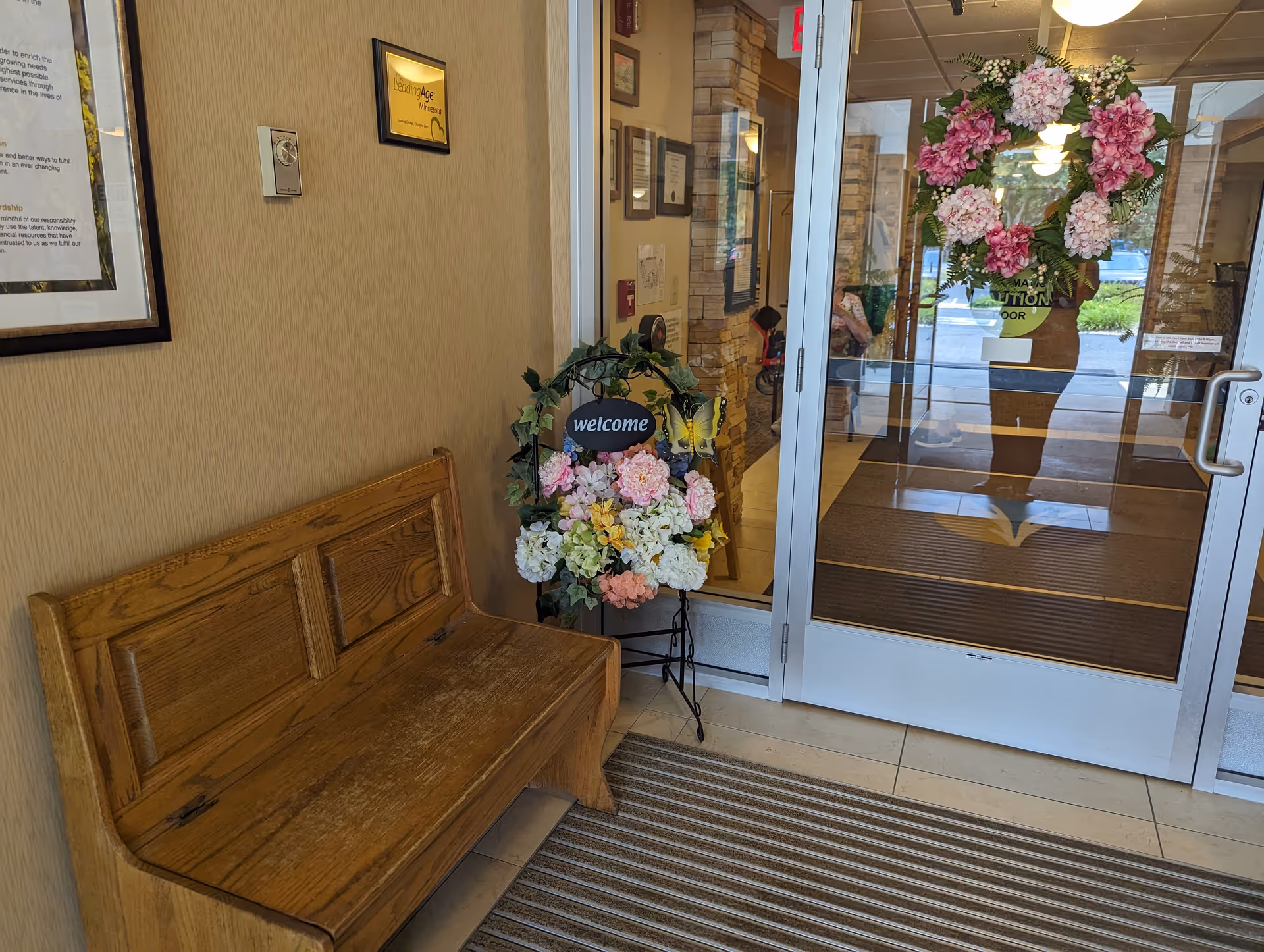 Entryway of a facility with a wooden bench on the left, a floral welcome arrangement on a stand, and glass double doors decorated with a pink and white flower wreath. The interior beyond the doors shows framed certificates and a stone accent wall.