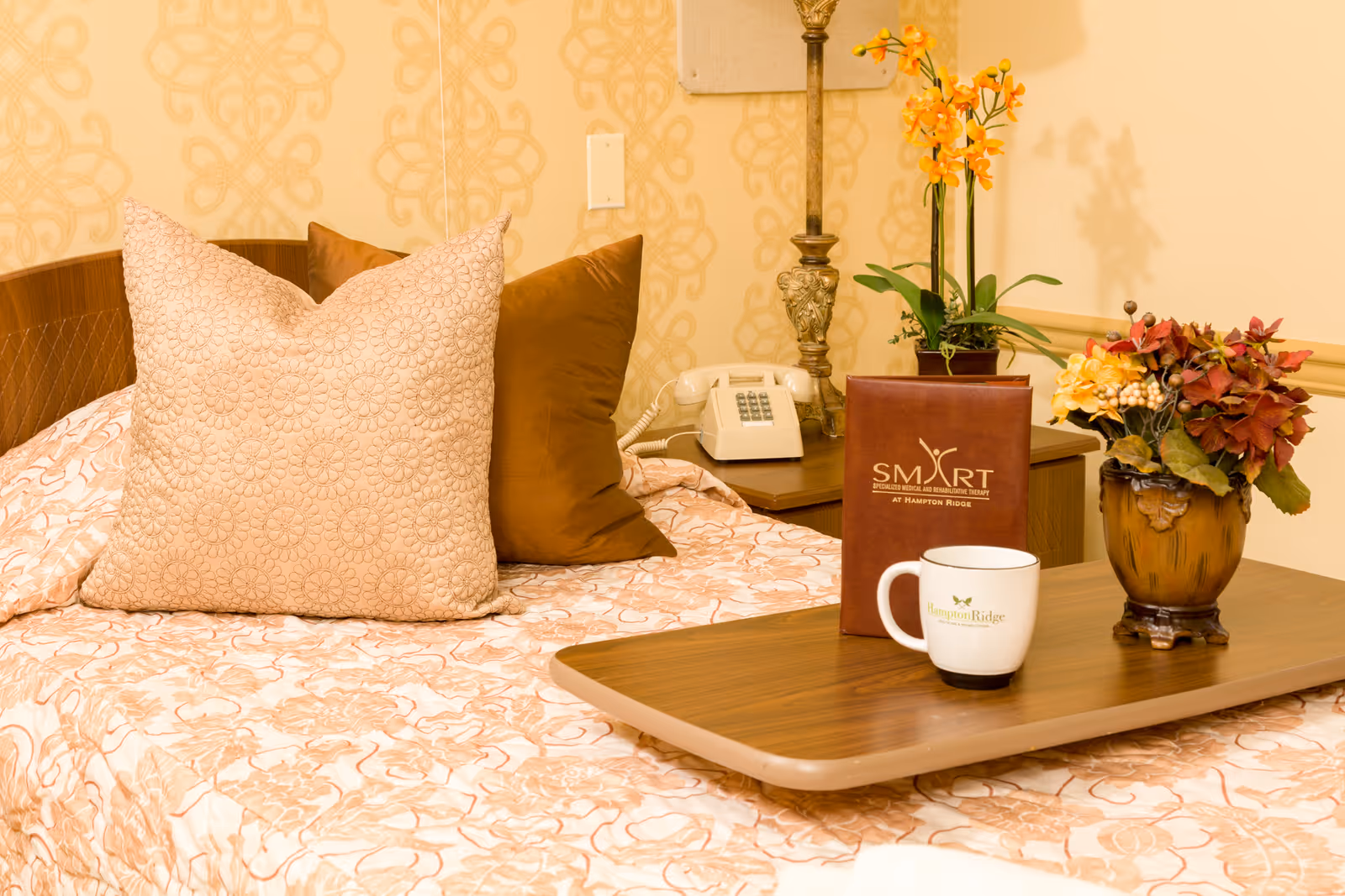 A neatly made bed with beige and brown pillows and a patterned bedspread. A wooden overbed table holds a white mug with Hampton Ridge logo, a brown folder labeled SMART at Hampton Ridge, and a vase with artificial flowers. A vintage telephone and a decorative lamp are on a nightstand beside the bed.