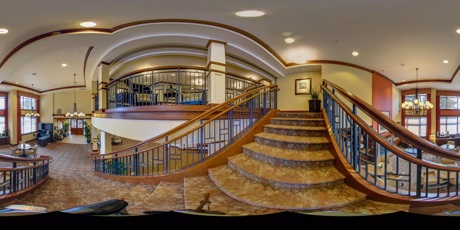 Spacious senior living facility interior with a carpeted curved staircase leading to a mezzanine and seating areas under chandeliers.