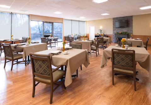 A dining room with several tables covered in beige tablecloths, each set with plates, cups, and small flower arrangements. Wooden chairs with patterned cushions surround the tables. Large windows with blinds allow natural light to fill the room. A stone fireplace with a mounted TV above it is visible on the far wall.