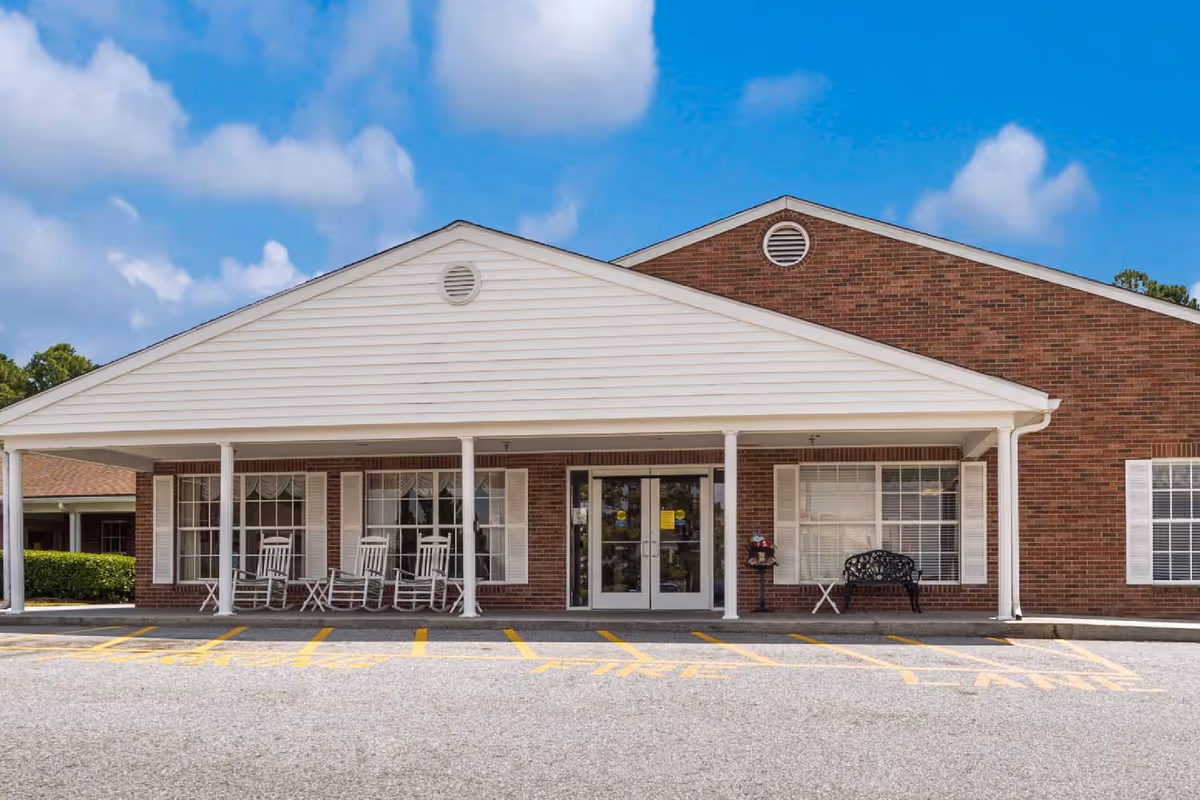 Front exterior view of Augusta Health Center showing a brick building with white trim, a covered entrance supported by white columns, rocking chairs and a bench on the porch, and a parking area with yellow painted lines in front.