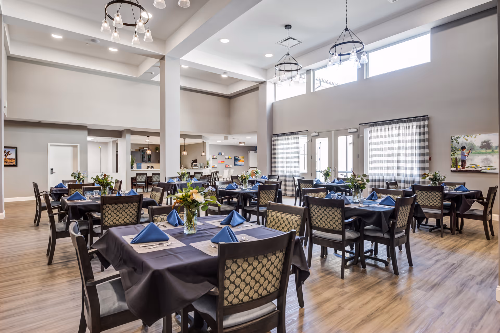 A spacious, well-lit dining room with multiple tables covered in dark tablecloths and set with blue napkins, silverware, and glassware. Each table has a floral centerpiece. The room features high ceilings with modern light fixtures, large windows with striped curtains, and light wood flooring. In the background, there is a counter area with bar stools and pendant lights.