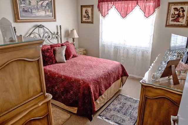 A cozy bedroom with a bed covered in a red patterned bedspread and matching pillows. The room features wooden furniture including a dresser and nightstand with a lamp. There are framed pictures on the walls and a window with sheer white curtains and a red valance letting in natural light. A small rug is placed on the carpeted floor near the bed.