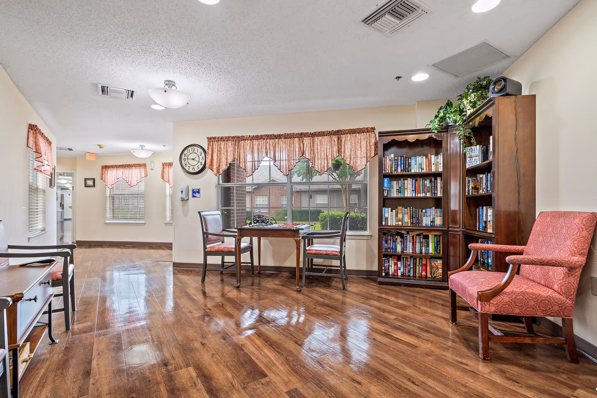 A bright and clean common area in a senior living facility with polished wooden floors, a small table with two chairs near a large window with red curtains, a wooden bookshelf filled with books, and a red upholstered armchair. The walls are light-colored, and there is a clock on the wall near the window.