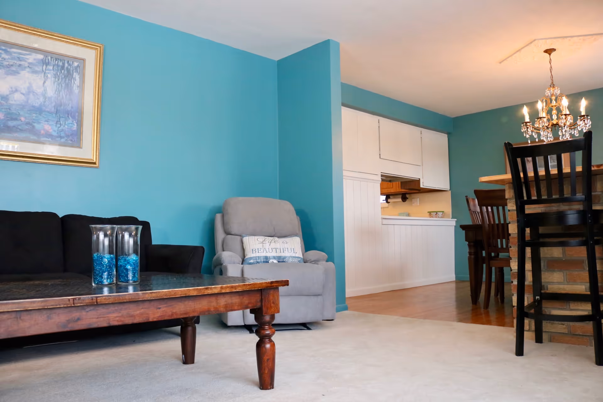 Interior view of a living room and dining area with teal walls. The living room has a black sofa, a gray recliner with a decorative pillow that says 'Life is Beautiful,' and a wooden coffee table with two glass vases filled with blue decorative stones. The dining area features a wooden dining table with chairs and a chandelier overhead. A pass-through window connects the dining area to the kitchen.