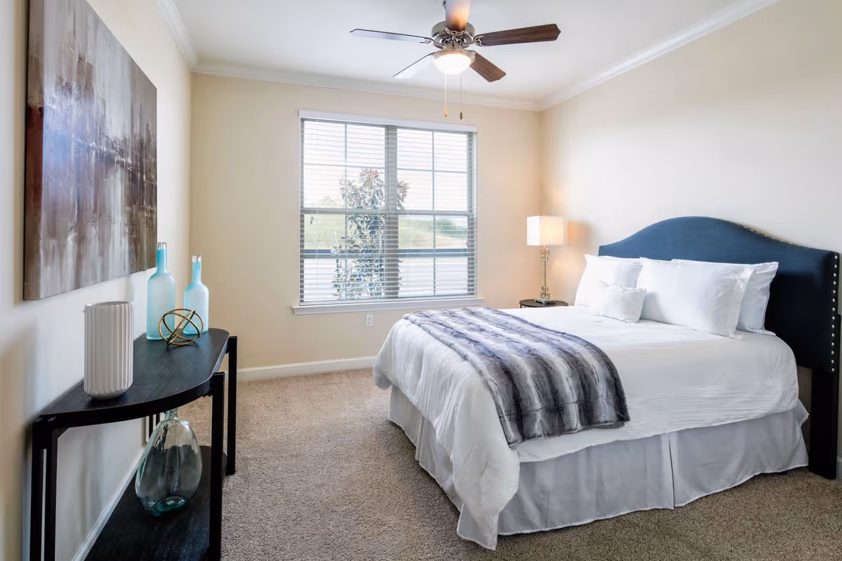 Bright, neatly staged bedroom with a bed, bedside lamp, ceiling fan, window, and a decorative console table with vases.