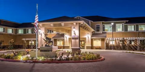 Night exterior view of Carlton Senior Living Elk Grove entrance with landscaping and flag.