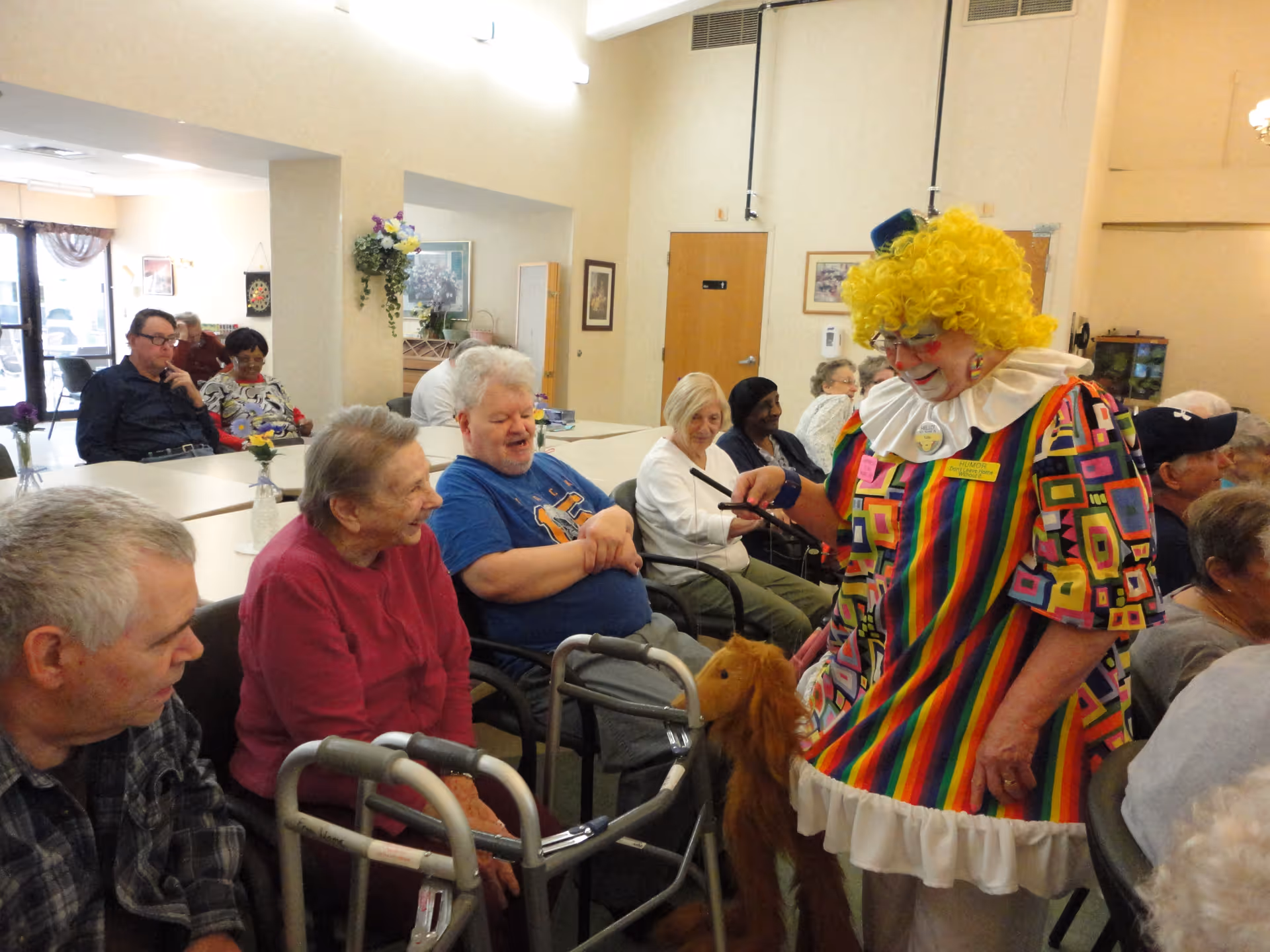 A group of elderly people seated in a common room, interacting and smiling with a person dressed as a colorful clown holding a puppet horse. The room has tables, chairs, and some wall decorations.