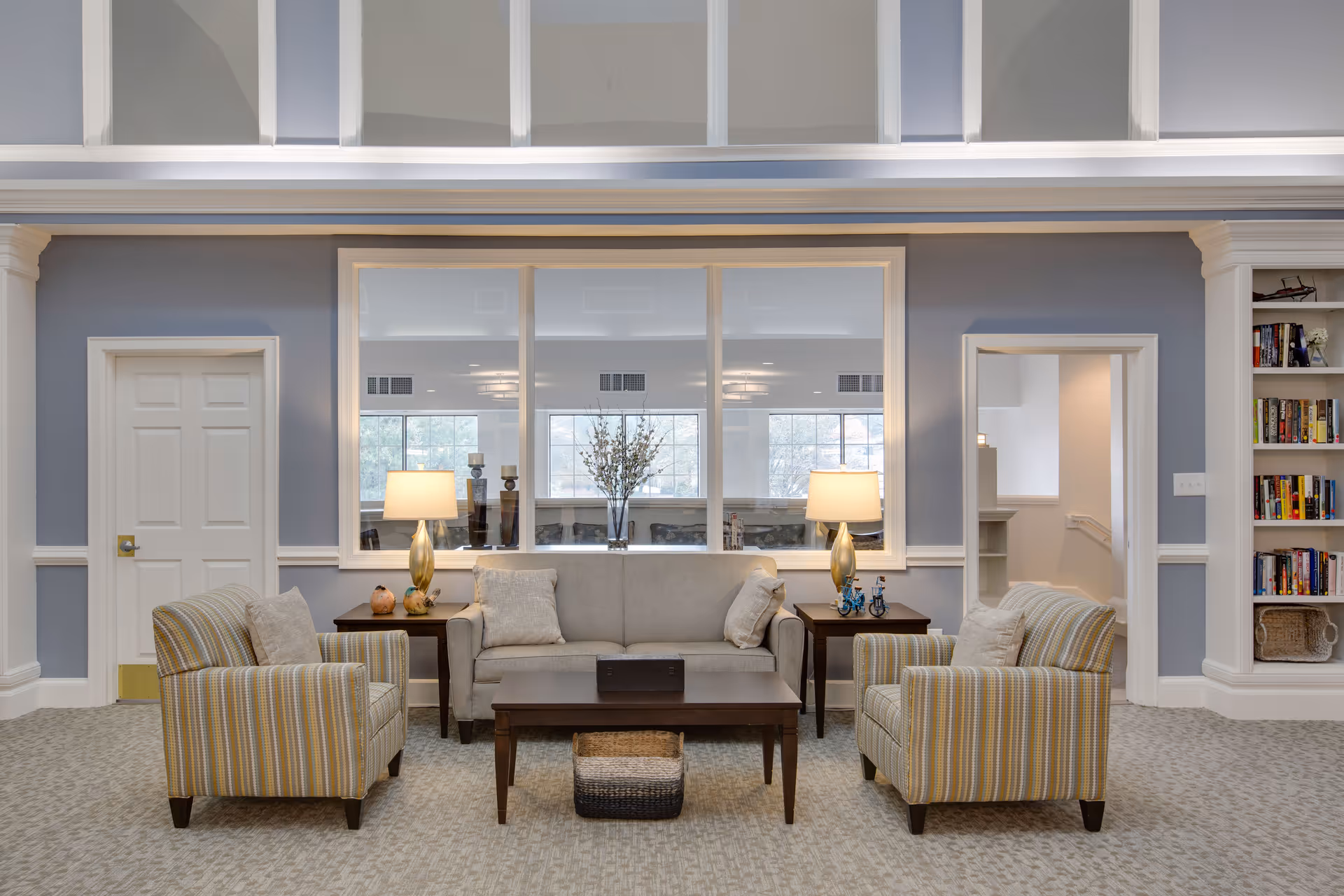 A cozy sitting area in a senior living facility with a beige sofa flanked by two striped armchairs. Two wooden side tables with lamps and decorative items are placed on either side of the sofa. A wooden coffee table with a basket underneath is in front of the sofa. The walls are painted light blue with white trim, and there is a large window behind the sofa reflecting another room with windows. Built-in bookshelves filled with books and baskets are visible on the right side.