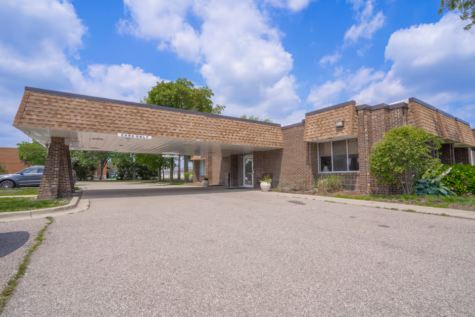 Front entrance of a low brick building with a covered drop-off canopy, driveway and parking area under a partly cloudy blue sky.