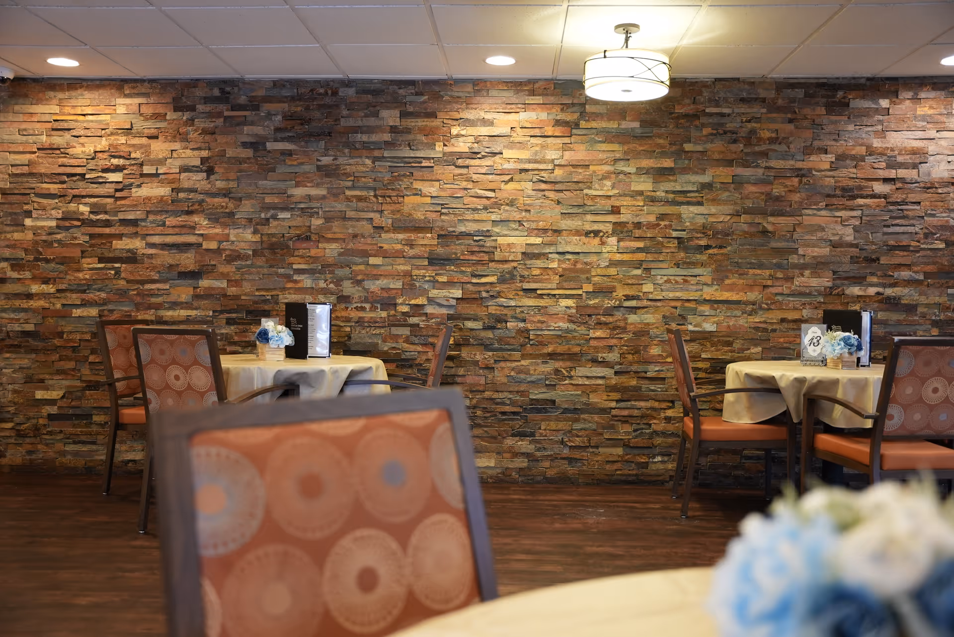 Dining room with round tables set with centerpieces, patterned chairs, and a stacked-stone accent wall under ceiling lights.