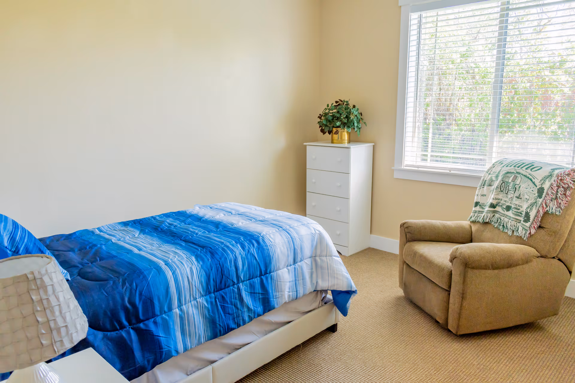 A bedroom with a single bed covered in a blue striped comforter, a white nightstand with a lamp, a white chest of drawers with a potted plant on top, a beige armchair with a green and white blanket draped over the back, and a window with white blinds letting in natural light.