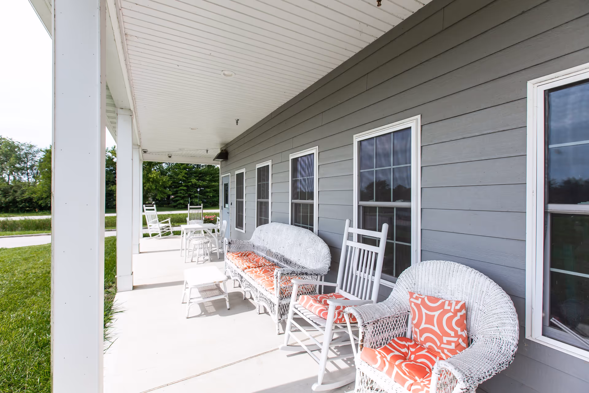 Covered outdoor porch area with white wicker furniture including a loveseat, rocking chair, armchair, and small tables, all with orange patterned cushions. The porch has a white ceiling and gray siding with multiple windows. Green grass and trees are visible in the background.