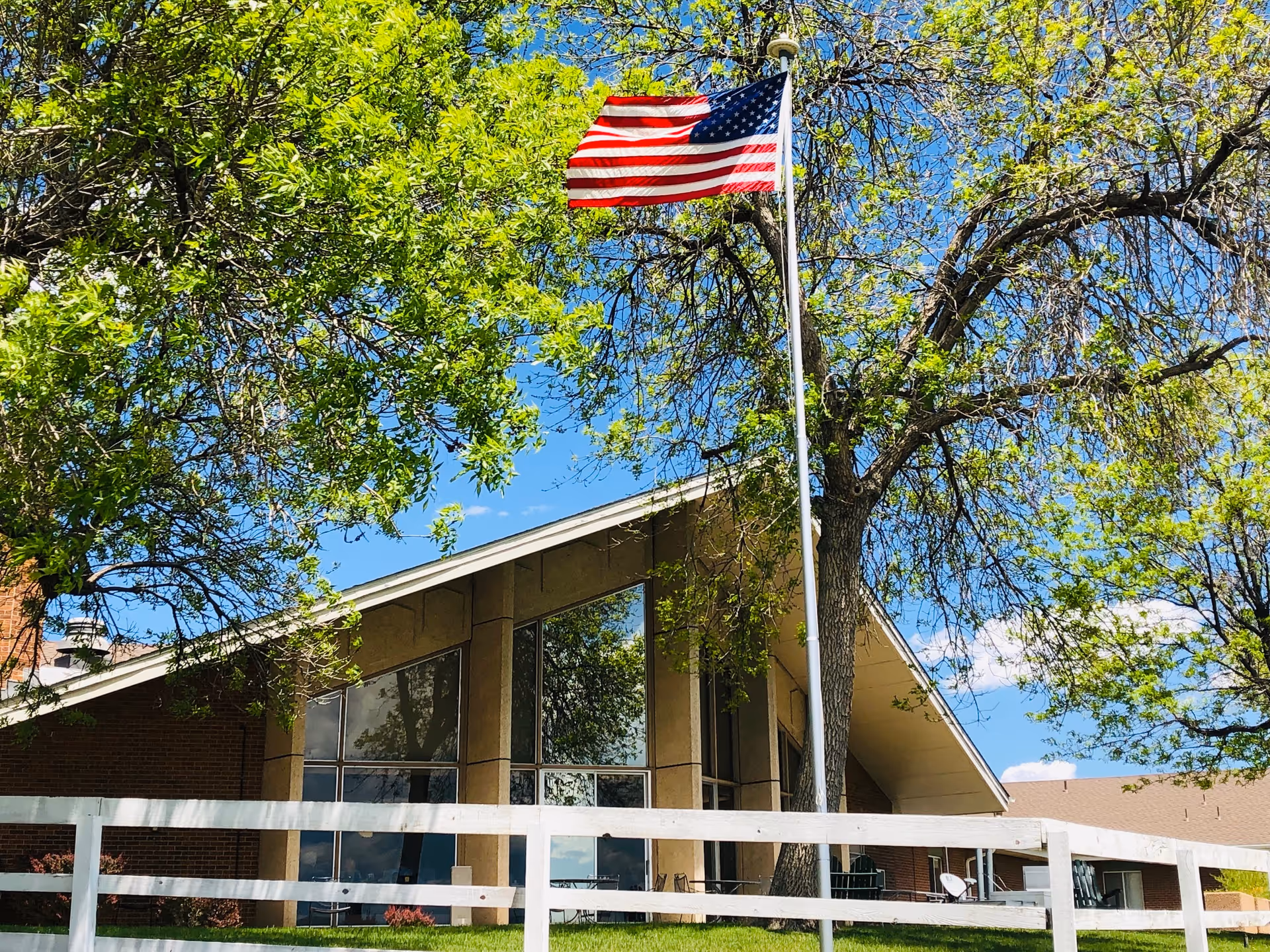 Exterior view of Mountain Vista Senior Living Community building with large windows and a slanted roof, surrounded by green trees and a white fence. An American flag is flying on a flagpole in front of the building under a clear blue sky.