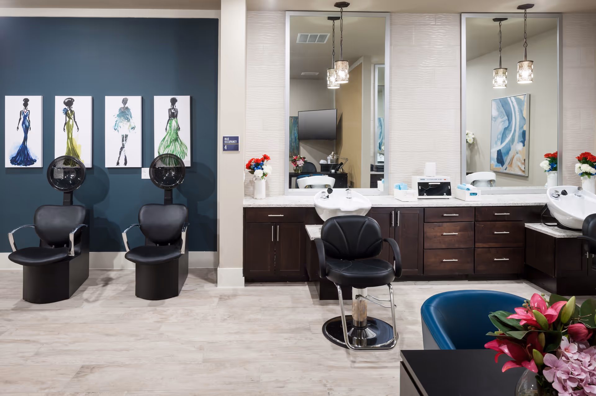 Interior view of a salon area with two black hair drying chairs against a dark teal wall adorned with four fashion-themed paintings. To the right, there are two large mirrors above a countertop with sinks and dark wooden cabinets underneath. A black salon chair is positioned in front of one sink. The room has light wood flooring and decorative flowers on the counter and table.