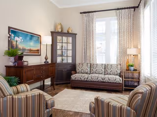 Sunlit living room with a patterned sofa and two striped armchairs arranged around a rug, wooden sideboard and glass-front cabinet by large curtained windows.
