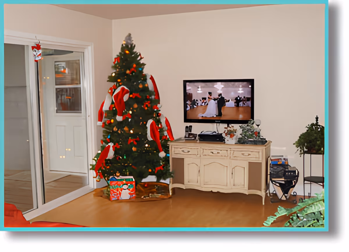 A living room decorated for Christmas with a Christmas tree adorned with red Santa hats and lights. There are wrapped presents under the tree. A flat-screen TV is mounted on the wall above a vintage-style cream-colored cabinet. The TV displays a scene of a bride and groom dancing. There are plants on the right side of the cabinet and a sliding glass door on the left side of the room.