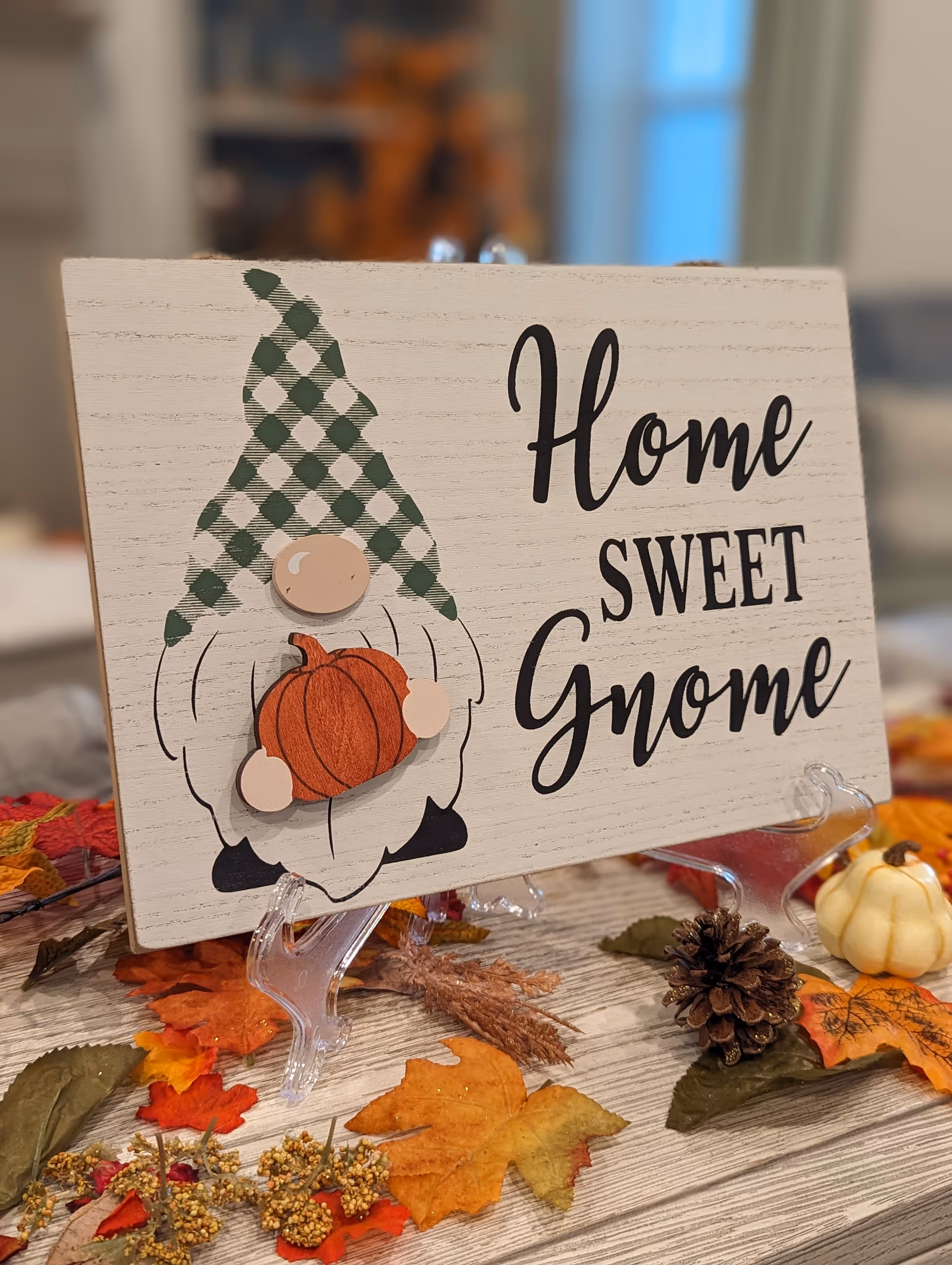 A decorative wooden sign with a gnome holding a pumpkin and the text 'Home Sweet Gnome' displayed on a clear stand. The sign is surrounded by autumn-themed decorations including colorful fall leaves, a pine cone, and small pumpkins on a wooden surface.